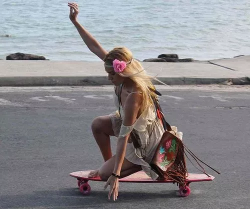 Девушка хиппи едет на скейте, у нее сумка украшенная бахромой и цветочный венок на голове