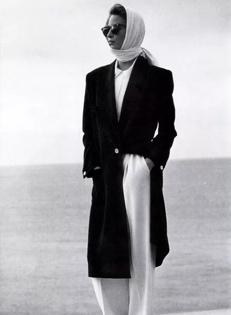 Модель в широких белых брюках и блузе, сверху черное длинное пальто