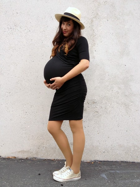Беременная девушка в обтягивающем черном платье, кеды и светлая шляпа