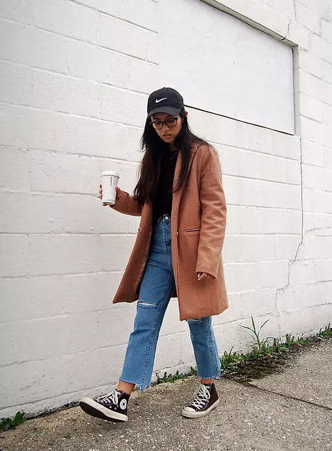 Девушка в рваных укороченных джинсах, коричневое пальто, кеды и кепка