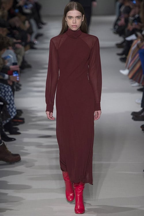 Модель в бордовом платье с рукавами и высоком воротником от Victoria Beckham