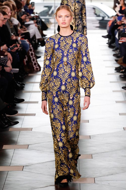 Модель в длинном синем платье с золотыми узорами и длинными рукавами от Tory Burch