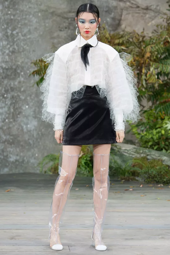 Модель в белой блузке с пышными руквами, черной мини юбке и прозрачные сапоги от Chanel