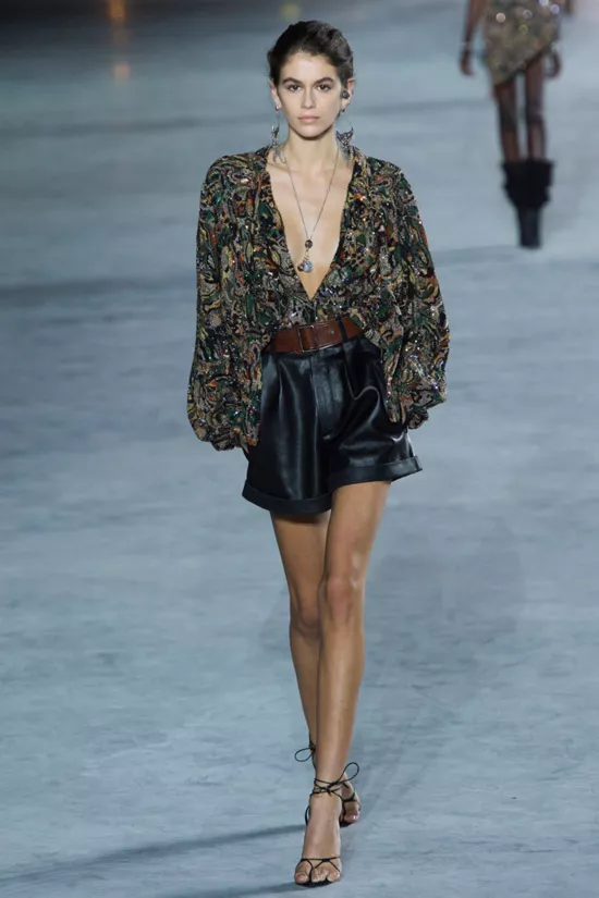Модель в темной блузке с длинными рукавами и кожаные шорты от Saint Laurent