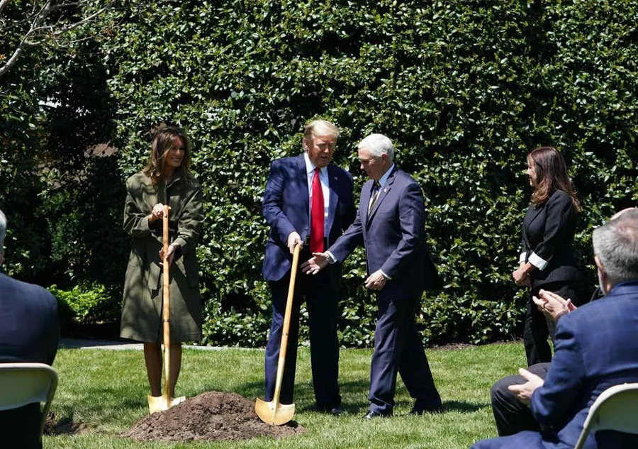 Мелания Трамп в оливковом плаще с поясом и лопатой в руках