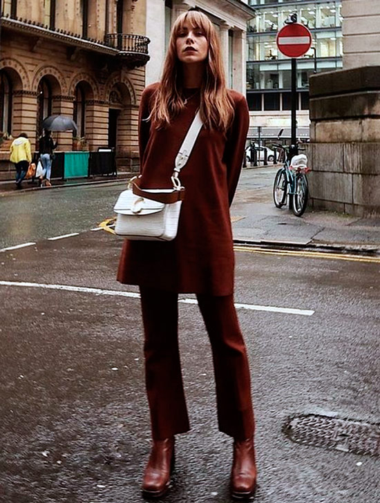 Девушка в коричневых брюках, удлиненный свитер и ботильоны, образ дополняет белая сумка