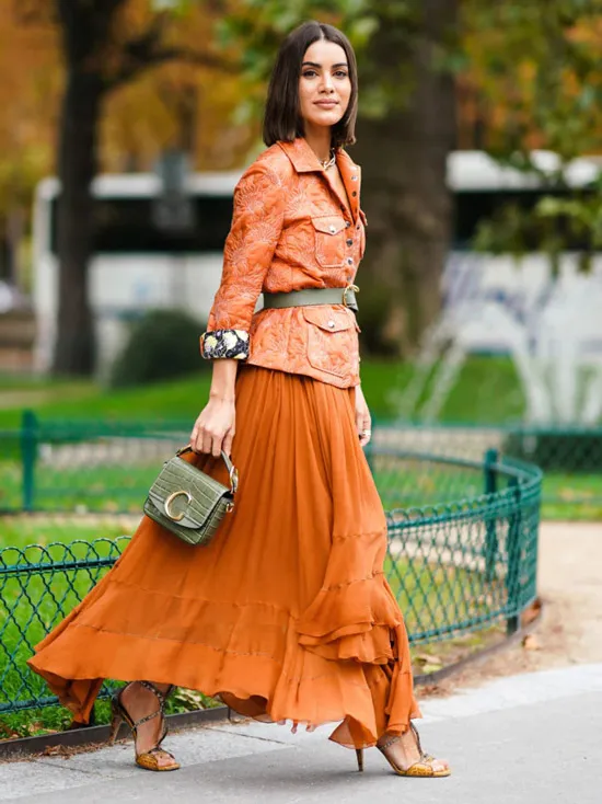 Девушка в воздушной юбке макси с воланами, оранжевый блейзер с ремнем на талии, образ дополняют босоножки на шпильке и серая мини сумочка