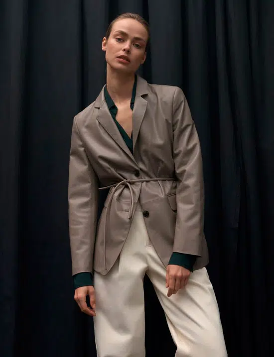 Модель в сером кожаном пиджаке с тонким поясом и белых брюках