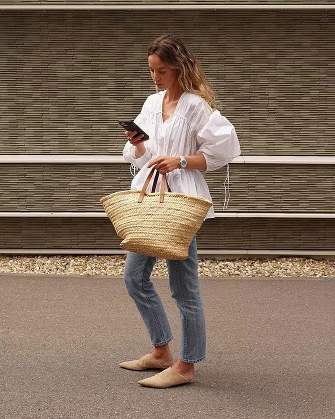 Девушка в прямых джинсах, белой свободной блузке и с соломенной сумкой