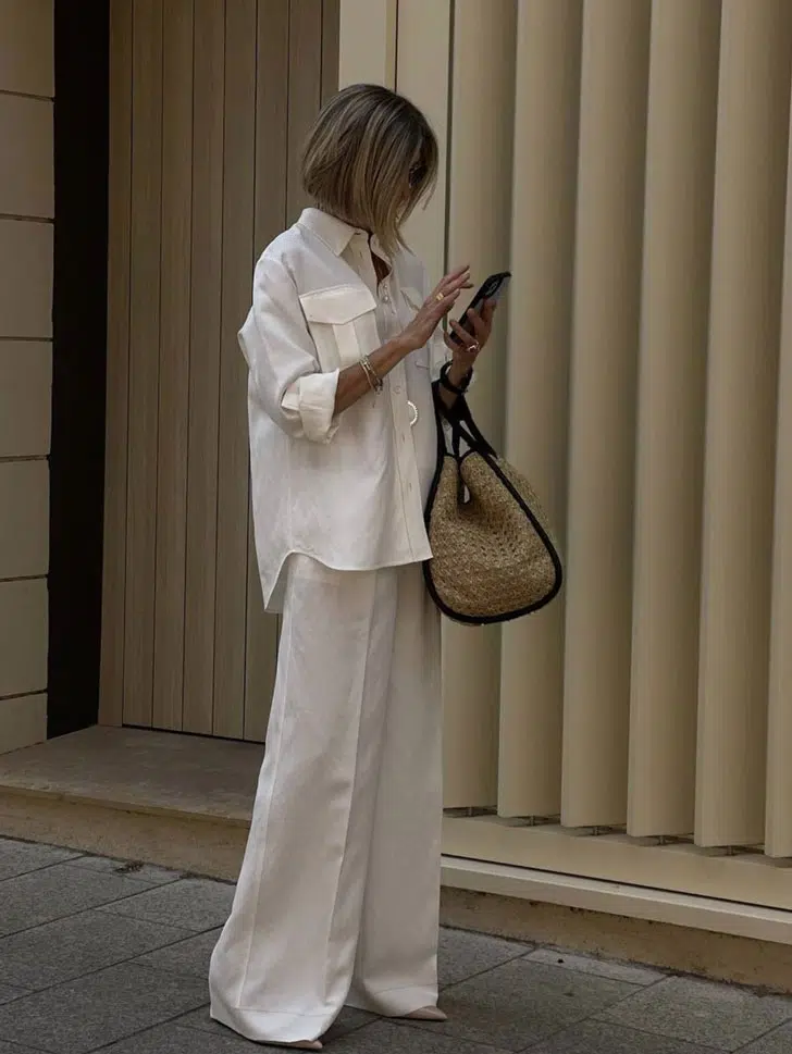 Женщина в белых широких брюках клеш, рубашке оверсайз и с соломенной сумкой