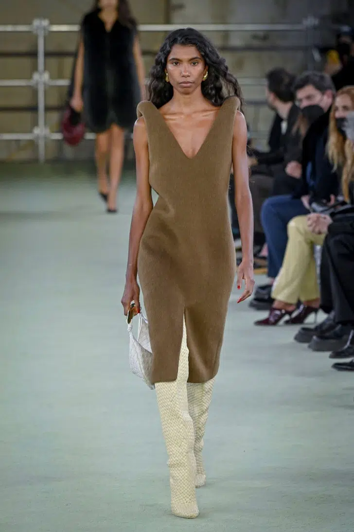 Модель в коричневом платье, высоких сапогах и с мини сумочкой