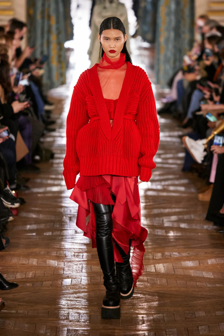 Модель в красном вязаном кардигане, легкой юбке с воланами и сапогах ботфортах на высокой подошве