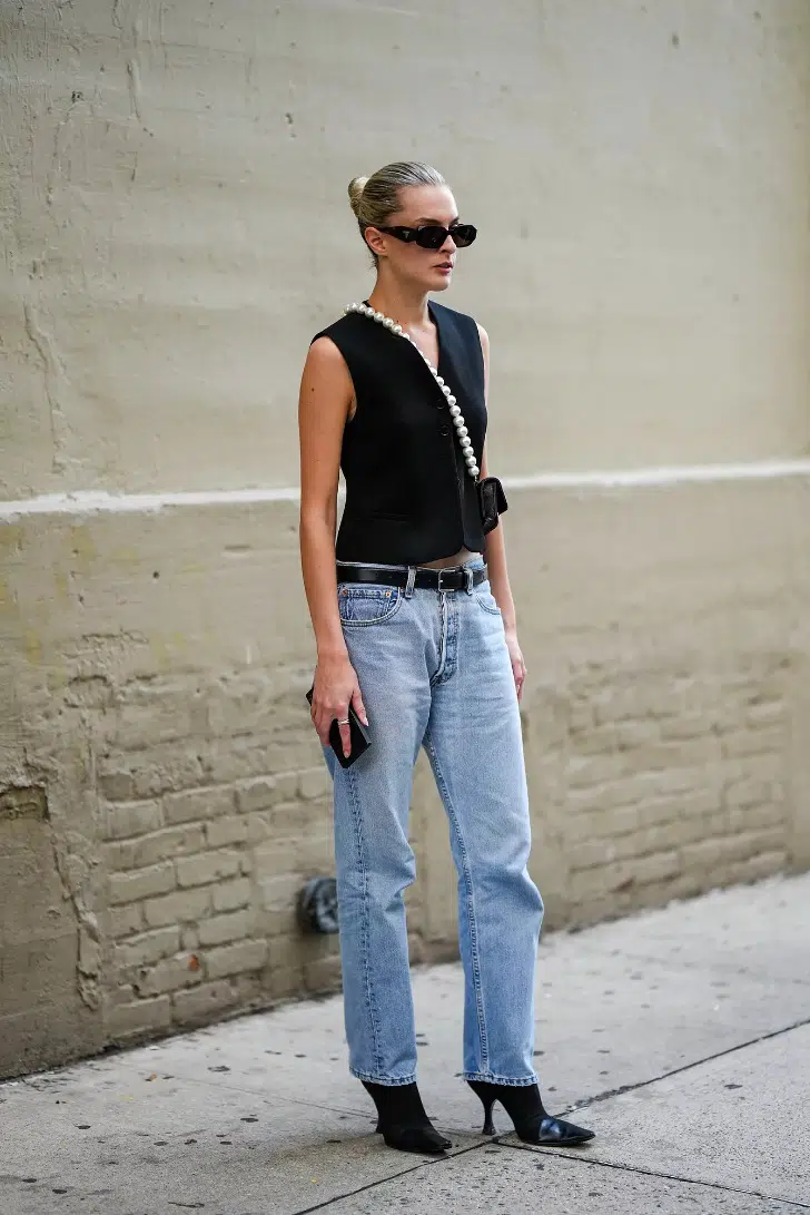 Девушка в прямых джинсах с ремнем, черной жилетке и ботильонах