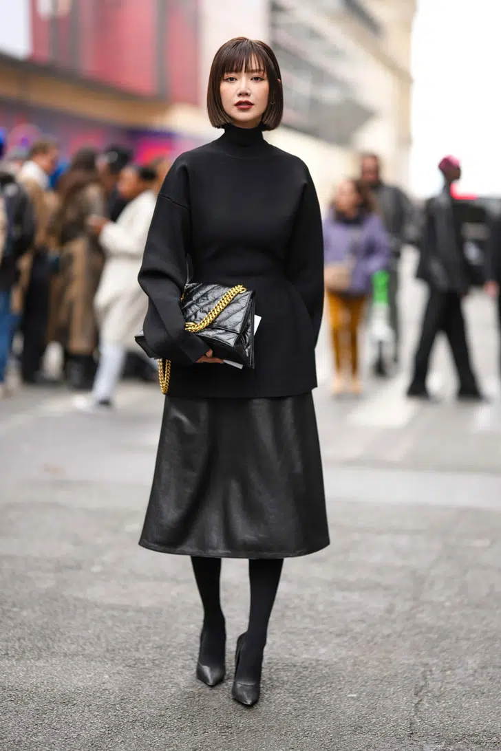 Девушка в черном образе с кожаной юбкой миди, удлиненной водолазке и стеганой сумкой на цепочке