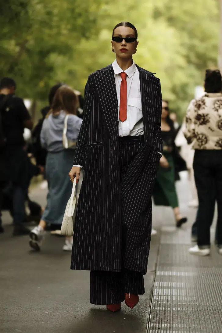 Девушка в длинном черном пальто в тонкую полоску, широких брюках и рубашке с галстуком