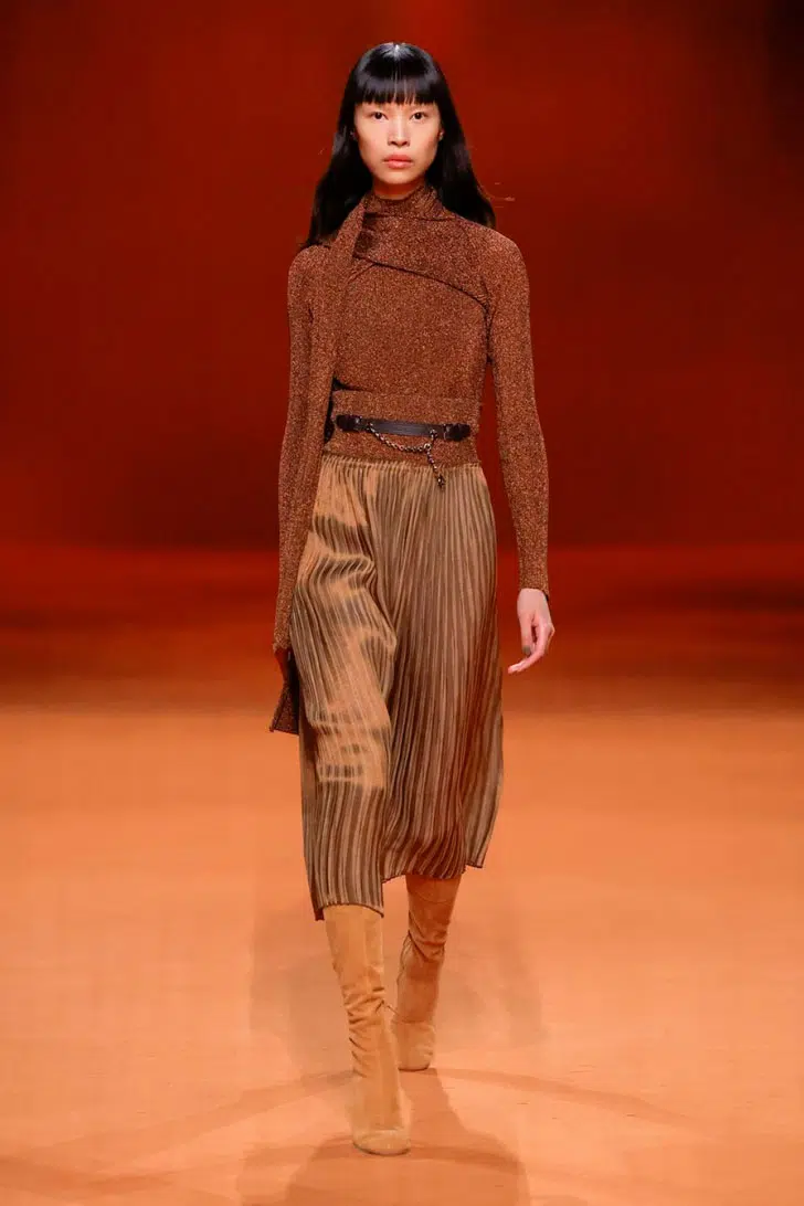Модель в плиссированной юбке миди, коричневом свитере и сапогах из замши от Hermès